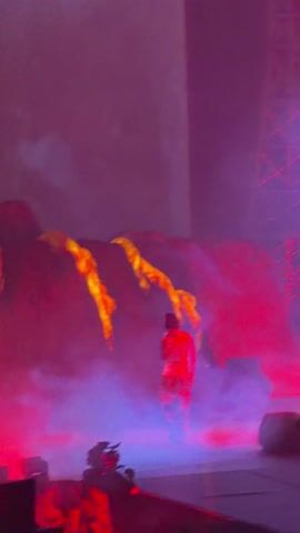 The video shows a stage lit with red and purple lights, with a performer in the center. The performer is wearing a white t-shirt, a red jacket, and camouflage pants. Behind the performer is a backdrop of what appears to be a large rock formation with yellow and orange designs on it, possibly representing lava or flames. A tall structure resembling an oil rig is visible in the background. There is a dense haze or fog on the stage, which combined with the colored lighting, creates a dramatic atmosphere. A camera operator is visible on the lower left of the stage, filming the performance.