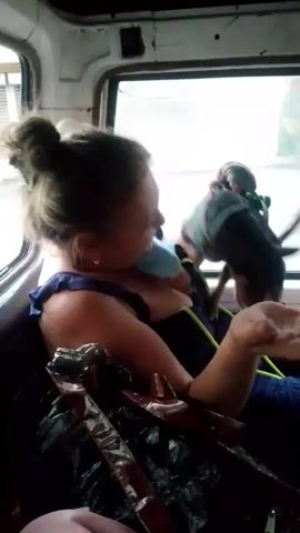 A woman with her hair pulled back in a bun and wearing a blue dress is seated in a vehicle. She is holding a small black dog on a leash. The dog is standing on her lap and looking away from the camera. The woman is smiling and looking at the dog. The vehicle appears to be an older model with a worn interior. There are plastic bags and other items in the foreground. Outside the window, there are buildings and a street scene, with a person walking in the distance. The lighting is dim, suggesting it might be overcast or nighttime.