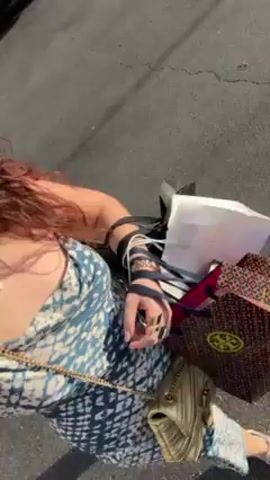 A person with long curly red-brown hair wearing a blue and white patterned sleeveless top and multiple bracelets on their left wrist is holding several shopping bags by their handles. The camera moves, revealing a street scene with buildings and cars in the background. The person appears to be standing in a parking area or street.