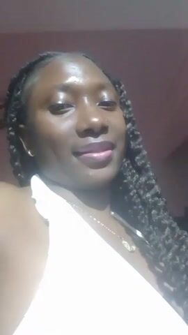 A medium-shot video features a dark-skinned woman looking directly at the camera. She has her hair styled in thick twists, and is wearing a white halter top and a gold necklace with a small charm. There is a blurry, possibly pinkish, background. The lighting is slightly harsh, causing some glare on her skin. The woman's expression is gentle, with a slight smile. There's loud music playing in the background.