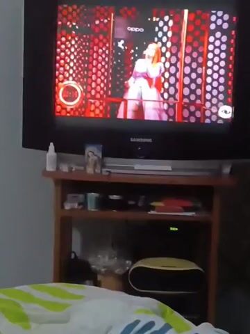 The video shows a Samsung TV on a wooden stand. The TV is playing a video game featuring a woman with a microphone singing against a red backdrop with white polka dots. The timer on the screen shows 0:12. On the stand below the TV, there are various items including a small bottle, a picture frame, containers, books, and an electronic device. The camera then pans down to a bed covered with colorful blankets and a white, fluffy pillow with a cartoon bear face.