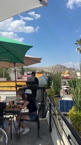 The video starts on a rooftop terrace with a DJ playing music and a woman sitting at a table. The camera then pans to show a church tower in the distance, surrounded by lush green bushes and palm trees. The sky is bright blue with scattered white clouds.