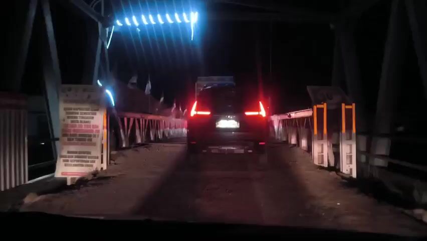 The video shows a dark-colored SUV driving slowly on a narrow bridge at night. The vehicle is heading towards the camera, with its brake lights illuminated. The bridge has metal support beams and a low barrier along the sides. There are also some decorations, possibly flags or lights, on the bridge's structure. The roadway appears to be unpaved or poorly maintained. The surrounding environment is very dark, with minimal ambient light. The camera is positioned from inside another vehicle, looking out through the windshield. A sign is visible on the left side of the bridge entrance.