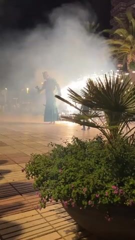 A person in a green costume stands in front of a pyrotechnic display. The display emits a bright shower of sparks and smoke. The scene appears to be outdoors at night, with palm trees and plants visible in the foreground. The ground is paved with tiles.
