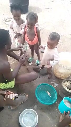 A video shows three young Black children, likely toddlers, standing in front of a dark-skinned adult man who is sitting on the ground. The child on the left wears a long-sleeved dress with light pink strawberries printed on it. The child in the middle wears a dark pink bathing suit and light green sandals, while the child on the right wears a long-sleeved white top. The man wears a bright green and black swim suit, and he is facing the children, interacting with them by gesturing with his hands. The ground appears to be dirt or sand. Several containers are on the ground, including a large clear plastic container filled with grains, a green plastic bowl, and a silver metal bowl. A black barrel is visible in the background.