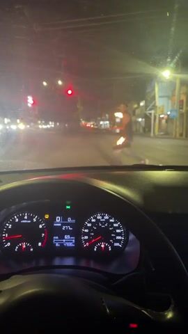 From the perspective of someone inside a car at night, a person juggling two flaming objects is seen in the middle of the street. The car is stopped at a red light. The person juggling is wearing a tank top and jeans. The surrounding environment appears to be a city street with buildings and other cars visible in the background. The dashboard of the car is visible in the foreground, showing the car is not in motion. The video is blurry due to the darkness and potential movement.
