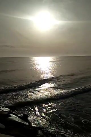 The video shows a sunlit ocean. The sun is bright and appears high in the sky, casting a shimmering reflection on the water. Gentle waves are seen approaching the shore. In the foreground, a dark, rocky edge is visible, possibly a pier or breakwater. The overall scene is one of a calm, natural environment. There are no people or specific actions being depicted.