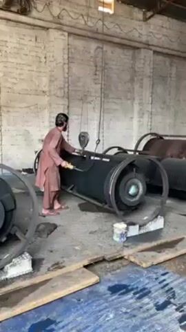 In an industrial setting, a man in a brown shalwar kameez and a black head covering is working with a black, cylindrical machine supported by a metal frame with wheels. The machine is suspended by a rope and pulley system. The man appears to be adjusting or operating the machine, using both hands. He is looking upwards at the pulley system. Several similar machines are visible in the background. The setting appears to be a warehouse or factory, with white brick walls, corrugated metal roofing, and a cement floor that appears wet in some areas. Paint cans sit on a block beneath the machine he’s working on. A dirty rag sits on the floor next to one of the machine’s wheels.