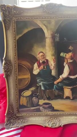 The video displays a framed painting depicting two men in a dimly lit interior, possibly a tavern or cellar. The man on the left, wearing a white shirt, red vest, and dark apron, is seated on a stool, holding a mug in his right hand and gesturing with his left. He appears to be speaking or explaining something. He has a reddish cap on his head and a smile on his face. The man on the right, also seated on a stool, is wearing a similar red vest and white shirt with a red cap. He is drinking from a green-colored mug or bottle. In the background, there are barrels, stone walls, a shelf with objects, and a hanging lantern. The floor appears to be made of stone or dirt. The painting is ornate, with a gilded frame exhibiting carved details. The video briefly pans across the painting, showing the entire scene and its frame. The overall mood suggests a moment of camaraderie and relaxation.
