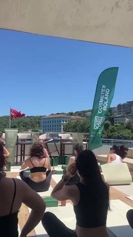 The video shows a group of people from behind, seated on yoga mats on a sunny day. The individuals are dressed in athletic wear, and some are actively participating in what appears to be a yoga or meditation session. In the background, there's a body of water with buildings and trees on the opposite shore, and a Turkish flag is visible flying from a pole. A large green banner with the text "COSMETEX ROLAND" and "CHANTECLAIRE MATCHA" is positioned to the right of the group. Several green bottles, likely cosmetic products, are placed in front of the participants. The overall atmosphere suggests an outdoor event or class taking place on a boat or a waterfront venue.