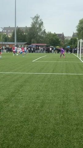 A soccer player in a red jersey slides on artificial turf to kick a soccer ball toward the goal. Another player in a purple jersey and a player in a blue jersey stand near the goal. The red jersey player successfully kicks the ball into the goal. Spectators cheer.