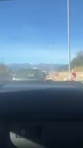 The video shows a driver's point-of-view while stuck in a traffic jam.  The road appears to be under construction, with orange and white construction barricades visible along the side of the road. There are several vehicles ahead, including a silver Volkswagen Passat with the license plate KO-BF-716. In the distance, there is a mountain range with some snow-capped peaks visible against a clear, blue sky. The dashboard of the car where the video is being taken is visible in the foreground. Music is playing.