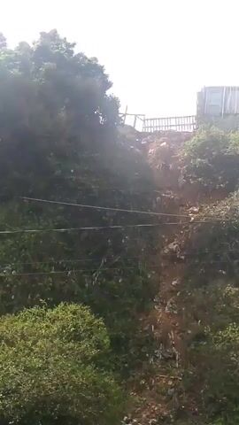 The video shows a landslide on a steep, vegetated slope. A partially constructed building is visible at the top of the slope. Debris from the landslide has moved down the slope, and some trash is scattered amongst the vegetation. A utility pole with wires is also present on the slope. The sky is overcast.