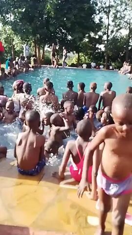 A group of approximately 40 children of various ages, mostly Black, are in and around a rectangular pool. Some are seated on the pool's edge, while others are standing or wading in the water. Splashing and general activity are evident among the children in the pool. Several adults, including a man in a red shirt, are present around the pool, seemingly supervising or observing the children. The pool is surrounded by a paved area and lush green trees. A small building is visible in the background. The scene suggests a recreational activity or a swimming lesson.