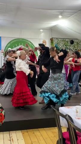 The video shows a group of people performing what looks like Flamenco dancing on a stage inside a tent.  They are dressed in colorful outfits including ruffled skirts, polka dots, and flowers in their hair.  Some men are present in shirts with polka dots. The stage is dark brown and elevated in the tent. The dance is taking place in a tent. One side of the tent wall is decorated with greenery and a logo. There are tables with covers and chairs visible in the foreground. The camera moves and pans to capture the dancers from various angles.