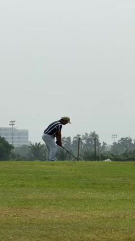 A man is shown on a golf course on a cloudy day. He is wearing a striped shirt, light-colored pants, and a green cap. He is in the process of swinging a golf club, with the club raised above his head. In the background, there are trees, some buildings, and what appear to be lights on poles. There is also a metal frame with some white material behind him. The man is positioned on a grassy area with another patch of grass in the foreground.