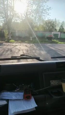 The video is filmed from inside a vehicle, looking out the windshield. The sun is shining brightly, creating lens flare and obscuring the view of the road ahead. Outside, trees and a building with garage doors are visible. The dashboard of the vehicle is in the foreground, with papers, keys, and a coffee cup on it. The overall impression is of a sunny day, possibly early morning or late afternoon, with the vehicle in motion.