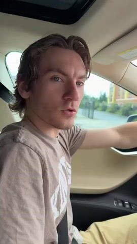 A young man with brown hair is sitting in the driver's seat of a car. He is wearing a beige t-shirt and tan pants. He is looking at the camera with his mouth open, appearing surprised or excited. The interior of the car is visible, including the dashboard and a sunroof. Outside the car, trees and buildings can be seen. The man is speaking Russian. He says he has a friend in the FBI and that he will find him in 3 minutes.