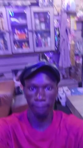 A person with dark skin is wearing a red t-shirt and a black cap. They are indoors, possibly in a store. Shelves stocked with various items are visible in the background. The lighting appears to have a purple hue. The person is looking directly at the camera.