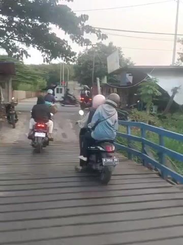The video displays a short clip of a street scene, likely in Southeast Asia, featuring multiple motorbikes and people. The camera appears to be mounted on a motorbike, suggesting a first-person perspective of someone riding or being a passenger. The road is paved with asphalt and some parts appear to be made of wooden planks, possibly indicating a bridge. Buildings and trees line the sides of the road, with some small shops visible. Several motorbikes are seen moving along the road, with people riding them. Two individuals are prominently featured in the foreground, riding a motorbike side-by-side. The person in front is wearing a light-colored jacket and a helmet, while the person behind is wearing a pink helmet and a jacket. They are both facing forward, observing the road ahead. The overall atmosphere seems to be that of a typical day with local traffic and everyday activities. The lighting suggests it might be daytime, possibly late afternoon or early morning given the soft light.