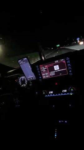 The video shows the interior of a car at night. The dashboard is illuminated with a digital display showing the time as 2:33 and a temperature of 23.0 degrees Celsius. A smartphone is mounted on the dashboard, displaying a navigation app. The car appears to be moving, as indicated by the blurred lights of other vehicles on a highway in the background. The infotainment screen is displaying music information.