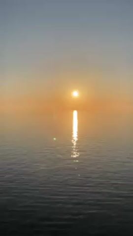 The video captures a serene scene of the sun setting over a calm body of water, likely an ocean or a large lake. The sun, appearing as a bright orb, dominates the horizon, casting a golden glow across the sky which transitions from orange near the horizon to a lighter blue overhead. The water's surface is relatively still, with gentle ripples reflecting the sun's light, creating a shimmering path towards the viewer. There are no visible people or objects in the scene other than the natural elements of the sky and water. The overall impression is one of peace and tranquility.