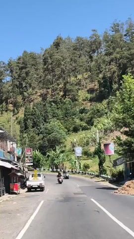 The video shows a road winding through a lush, green, mountainous area on a sunny day. The road is paved, and there is a white truck parked on the left side. Several motorcycles and a silver car are driving on the road. There are trees and vegetation on the hillsides. Billboards are visible along the road, and there are some small shops or stalls on the left. The sky is clear and blue.