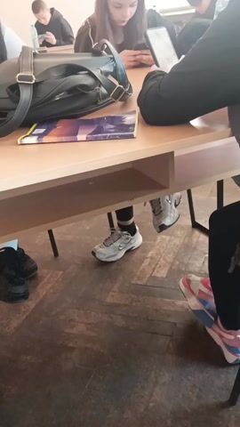 The video shows a classroom scene. Several students are sitting at desks, using their phones.  A black bag and a book are on the desk in the foreground. Below the desk, the feet of several students are visible. They are wearing different types of sneakers. The floor is wooden.  Someone shakes the table slightly, and a student gasps.