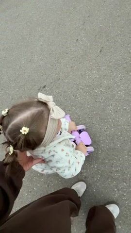 A person walks along an asphalt path while holding a small toddler who sits on a purple push toy. The toddler has brown hair pulled back into two small pigtails, each secured with a yellow flower clip. The toddler wears a beige headband with a bow, and a floral patterned jacket. The person holding the toddler is wearing a brown sweatshirt and matching pants, with white sneakers. The scene is set outdoors, showing trees along the edge of the path.