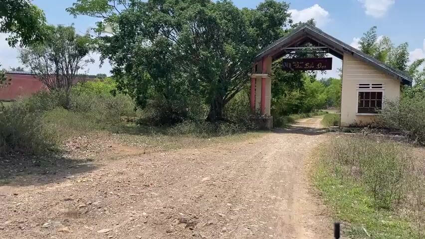 The video shows a dirt road leading through an entrance gate. The gate has a sign above it with writing on it, but the writing is difficult to read clearly. The gate is flanked by trees and bushes. There is a small building next to the gate on the right. The road continues into a wooded area. The sky is blue with some clouds.