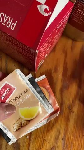 The video shows a close-up view of a person's hand interacting with tea bags. The hand is holding a tea bag with an image of lemon and ginger and text that appears to be in a foreign language. The person then places the tea bag into a red and white cardboard box. Inside the box, other tea bags are visible. The background is a wooden surface. The video is shot in a vertical orientation.
