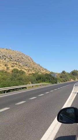 The video shows a view from a car traveling on a paved road. The road has white lane markings, including directional arrows. On the left side of the road, there is a rocky, sparsely vegetated hillside with some scattered trees. The sky is clear and blue. In the distance, on top of the hill, there are what appear to be communication towers or antennas. The right side of the video shows the car's side mirror and a portion of the passenger side door. The scene suggests a scenic drive, likely in a Mediterranean or similar climate.