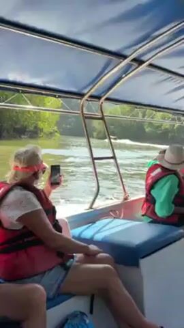 The video depicts a boat ride. Several passengers are visible, all wearing life jackets. One passenger is using a phone to take a photo or video. The boat is moving on a river or other body of water, with green foliage lining the banks. The boat is covered by a blue canopy. The day appears to be sunny.