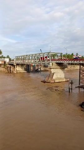 The video shows a view of a steel truss bridge spanning a wide, muddy river. The river appears to be high, possibly due to recent rain or flooding. The bridge is decorated with Indonesian flags along its sides. There are concrete supports in the river underneath the bridge, and some wooden planks. There's a crane on the bridge. The sky is overcast.