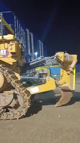 A Caterpillar bulldozer is parked outdoors at night. The machine is yellow with black tracks and has a large blade. The tracks are visible, as is the side of the bulldozer. There is a ladder attached to the side of the bulldozer for access. In the background, there are industrial pipes and lights. Another piece of heavy machinery, possibly a smaller excavator, is partially visible to the right of the bulldozer. Further in the background, a blue building or wall is illuminated by bright lights. There are also gas cylinders and welding equipment in the middle ground. A person appears to be working on a piece of equipment that is propped up on blocks, and sparks are visible, suggesting welding or grinding. The ground is dirt or gravel.