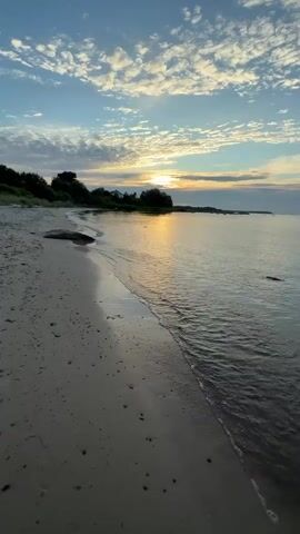 A still shot of a beach at sunset. The camera is positioned at the edge of the water, looking out towards the ocean. The sky is a pale blue with scattered white clouds. The sun is setting on the horizon, casting a golden glow on the water and the clouds. The beach is made of sand and pebbles. There are some trees and bushes on the left side of the frame. The waves are gently lapping at the shore. The overall mood is peaceful and serene.
