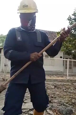 A person wearing a yellow hard hat, a dark blue long-sleeved shirt, dark blue pants, orange rubber boots, and a red face mask is sweeping with a broom. The person is standing on a cracked brick path in front of a building with a tiled roof. There are rocks and debris on the ground to the right of the person.