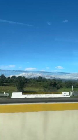 A clear blue sky dominates the top half of the frame. Below the sky, a chain-link fence runs horizontally across the middle of the image. Beyond the fence, a landscape of green fields and scattered trees unfolds. A stone wall with white posts is visible behind the fence, and further in the distance, a hazy mountain range can be seen. In the foreground, a bright yellow stripe runs along the bottom edge of the frame. The image appears to be taken from a moving vehicle, indicated by reflections on the glass and the slight blur of the scene.
