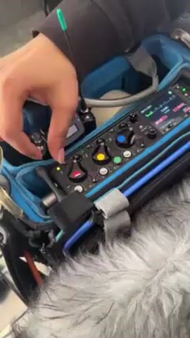 Here's a description of the video:

A person's hand adjusts a knob on a piece of audio equipment inside a blue carrying case. The equipment has various knobs, buttons, and a small screen displaying audio levels. A wireless microphone receiver is also visible in the case, along with a fuzzy microphone windscreen.