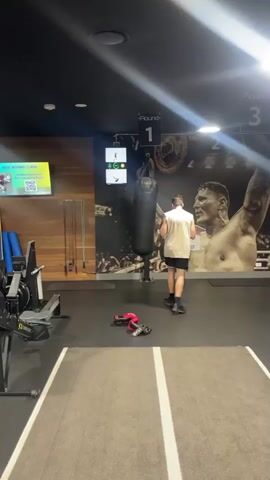 A man in a white tank top and black shorts is punching a black boxing bag in a gym. A scoreboard hangs above the boxing bag, displaying "Round 1". Two red boxing gloves and a pair of shoes are on the floor in front of the bag. To the left of the boxing bag is a rowing machine. The gym has a dark ceiling with spotlights and wooden paneling on the wall. A large mural of a boxer is visible on the wall behind the boxing bag. A screen on the left wall displays "BOXING CLASS".