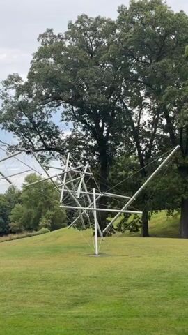 The video shows a long shot of a large outdoor sculpture on a grassy hill. The sculpture is made of silver metal rods and appears to be abstract, with many intersecting lines and shapes. Behind the sculpture, there are large trees with green leaves, and the sky is cloudy. The camera is panning slowly from left to right, providing a wide view of the sculpture and its surroundings. There are no people visible in the video. The grass is well-maintained and appears to be a typical park or garden setting. The lighting suggests it is daytime, possibly overcast.