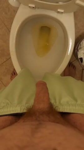 The video presents a first-person perspective of a man standing in front of a toilet with his pants down. The toilet contains yellow urine. His bare feet are visible on either side. A small metal waste bin with tissue paper is next to the toilet. The bathroom has a tan tile floor.
