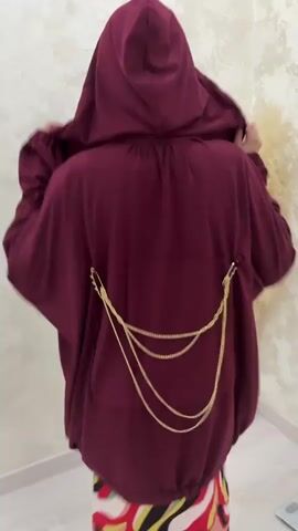 This media file shows the back of a person wearing a dark red hooded jacket with a drawstring hem. The jacket has three gold chains draped across the back, attached at each side with gold clasps. The person is standing in front of a light-colored wall with a textured pattern. To the right of the person, a large, fluffy, light-colored decorative item is visible. The lower half of the person's body is visible, wearing a brightly colored skirt or dress with a swirling pattern of red, yellow, black, and white. The overall impression is of a fashion showcase or a demonstration of clothing. The lighting appears to be artificial, suggesting an indoor setting. There is no visible text in the media file.