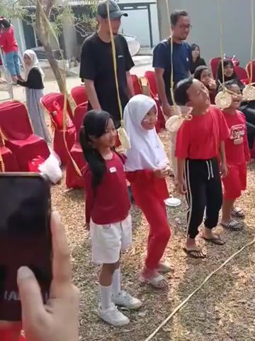 The video shows an outdoor game with children participating. They are tied to strings and have crackers attached to their faces. The children are trying to eat the crackers without using their hands. Adults are watching and some appear to be supervising. The environment appears to be a yard or a park with trees and some chairs visible.