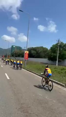 The video shows a group of cyclists riding on a paved road on a sunny day. The cyclists are wearing matching yellow, red, and blue jerseys. They are riding in a tight formation, with one cyclist ahead of the rest. The road is smooth, with a white line marking the edge of the lane. On the right side of the road, there is a grassy area with trees and a white wall. In the distance, a building and a mountain can be seen. There are light poles and power lines along the side of the road. There is also a red and yellow advertisement. The camera appears to be moving along with the cyclists, possibly from a vehicle.