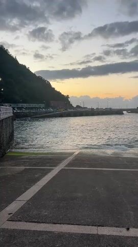 The video shows a body of water with a rocky, forested hill on the left. In the distance, there is a pier with lights and a bridge. The sky is cloudy with a hint of sunset. The foreground is a concrete surface with lines marked on it. There are cars parked in the distance near the hill. The water is slightly choppy.