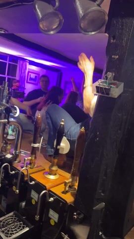 The video shows a woman dancing on the bar in a pub. The bar is made of wood and has several beer taps. The pub has several patrons and is lit with blue and red lights.