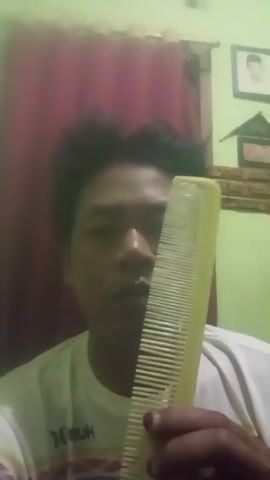 A medium shot shows a person's face as they comb their hair with a yellow comb. The person is wearing a white t-shirt with a dark stripe on the shoulder. The background is a room with a burgundy curtain and a framed picture on the wall. The lighting is dim.