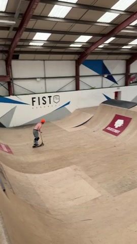 The video shows an indoor skate park. The walls are white with blue and grey geometric designs. On the left wall the word “FIST” is written in bold black letters, followed by the word “Adventurous”.  The ceiling is high, with exposed steel beams and skylight panels. The floor is made of wood, with various ramps and inclines for skating. A person wearing an orange helmet and grey pants is on a scooter on one of the wooden ramps. They perform a backflip on the scooter, landing back on the ramp, but appear to fall while riding down the ramp. A large red sign with the word “SKATEHUT” in white lettering with a house design is visible on the ramp.