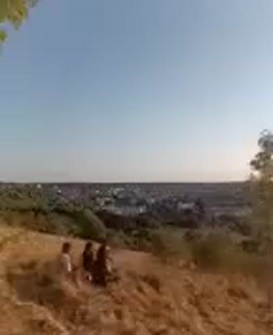 A video clip shows a group of people gathered on a grassy hill overlooking a town. The sun is setting, casting a warm glow over the landscape. The people appear to be enjoying the view and each other's company. One person is seen taking a picture of the sunset. The overall atmosphere is one of peace and relaxation.
